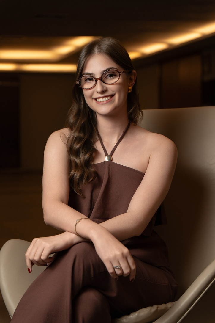 Ana Tamanini sorrindo, sentada em uma poltrona clara, de óculos e vestido marrom