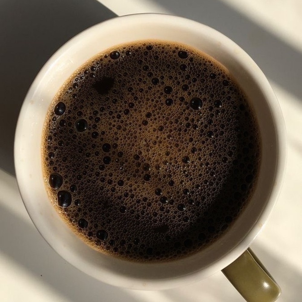 Xícara de café preto vista de cima, sobre uma mesa clara com sombras de janela